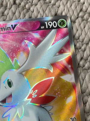 Pokémonkort Shaymin V holografisk - Säljer ett Pokémonkort med Shaymin V-motiv. Kortet är holografiskt med färger i silver, grönt, rött och rosa. HP 190 och grästyp-symbol syns i hörnet. Baksidan har klassiskt blått Pokémon-tryck med Pokéboll. Perfekt för samlare eller Pokémonfans.