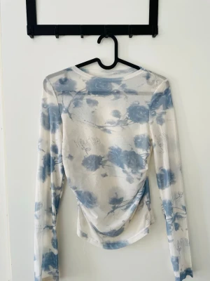 Blommig mesh topp med lång ärm - Säljer en transparent mesh topp med blått blommönster på vit botten. Går fint att använda som vanlig top eller över bikini.