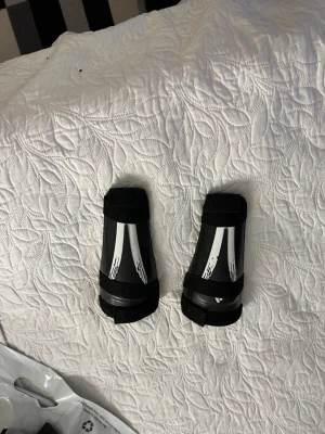 Svarta benskydd för fotboll  - Säljer ett par svarta benskydd för fotboll med vita detaljer och kardborreband. Skydden är formade för att sitta stadigt runt benen och har en hård yta för extra skydd. Perfekta för träning.