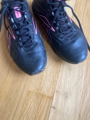 Svarta Reebok skor  - Snygga svarta gympaskor från Reebok med rosa detaljer och logga på sidan. Perfekta för en sportig och trendig look.