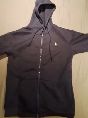 Mörkblå hoodie från Polo Ralph Lauren - Snygg mörkblå hoodie med dragkedja från Polo Ralph Lauren. Klassisk design med huva och dragsnören, samt fickor framtill. Ikoniska vita loggan broderad på bröstet. Perfekt för en chill och stilren look.