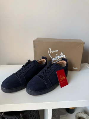 Mörkblåa sneakers från Christian Louboutin - Snygga mörkblåa sneakers från Christian Louboutin i mocka med ton-i-ton broderad logga på hälen. Klassisk låg modell med mörkblå snörning. Perfekta för dig som vill ha en stilren och exklusiv look.