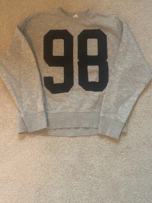 Grå sweatshirt med tryck från H&M - Grå sweatshirt från H&M Divided med stort svart '98'-tryck på framsidan. Tröjan har rund halsringning, ribbade muddar och är gjord i mjukt material med borstad insida för extra komfort.