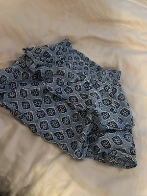 Blå kortkjol med mönster Gina Tricot - Kortkjol från Gina Tricot i ljusblått med grafiskt mönster i vitt och mörkblått. Kjolen har elastisk midja och är gjord i ett lätt, luftigt material som passar perfekt till sommaren. Snygg loose passform för en avslappnad look.