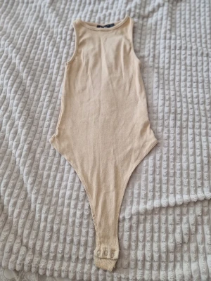Beige ribbad body från Boohoo - Säljer en beige ribbad body från Boohoo med rund halsringning och ärmlös design. Materialet är stretchigt och mjukt, perfekt för att styla med jeans eller kjol. Praktisk knäppning i grenen för enkel på- och avtagning.