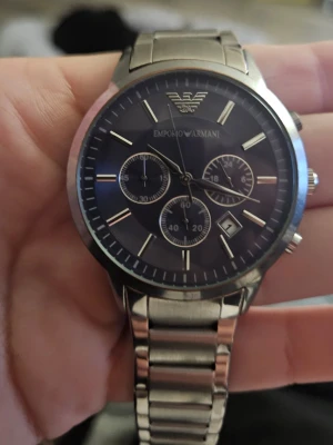 Emporio Armani silverfärgad klocka - Stilren klocka från Emporio Armani med silverfärgad länkarmband i metall. Svart urtavla med tre mindre subdials, datumvisning och tydliga indexmarkeringar. Klassisk design med logotypen högst upp på urtavlan och smidig viklås på armbandet. Köpt på Vinted, vet inget om den. Pli*k skapade beskrivning