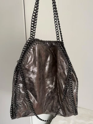 Stella McCartney Falabella liknande handväska - Säljer en metallicgrå Stella McCartney Falabella liknande handväska med kedjedetaljer runt kanterna och som axelrem.