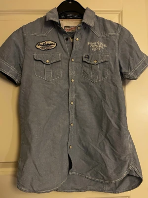 Blå jeansskjorta Petrol Industries stl 164 - Blå jeansskjorta från Petrol Industries med korta ärmar, tryckknappar och två bröstfickor med lock. Skjortan har vita kontrastsömmar, broderade detaljer och patch på bröstet. Stl 164 