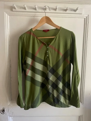 Vintage Burberry Henley  - Säljer en grön långärmad topp från Burberry med klassiskt rutigt mönster i svart och beige. Toppen har knappar framtill och är gjord i mjuk bomull. Perfekt för dig som gillar stilrena och ikoniska plagg.