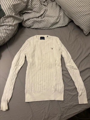 Vit kabelstickad tröja från Gant - Säljer en klassisk vit kabelstickad tröja från Gant med rund halsringning och diskret logga broderad på bröstet. Tröjan har långa ärmar och ribbade muddar vid ärmslut och nederkant. Perfekt för en stilren och tidlös look.