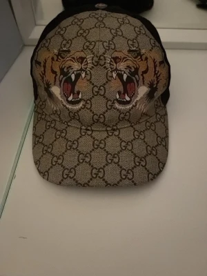Gucci keps med tigertryck - Säljer en snygg äkta  Gucci keps med klassiskt GG-mönster i beige och brunt. Framtill pryds kepsen av två detaljerade tigerhuvuden med öppen mun. Skärmen är böjd och baksidan är svart. Perfekt för dig som vill sticka ut med en exklusiv accessoar. Köpt ifrån Gucci i Italien 
