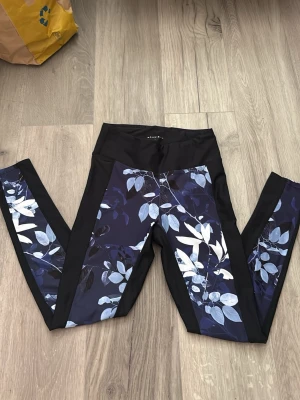Blommiga leggings från Röhnisch - Snygga leggings från Röhnisch med svart bas och blått blommönster längs benen. Hög midja och stretchigt material som sitter skönt. Perfekta för träning. Knappast använda 