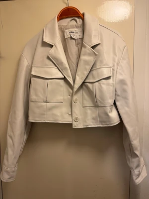 Vit cropped skinnjacka  - Snygg vit cropped skinnjacka (skinnimitation)  med två stora fickor framtill och klassisk krage. Jackan har raka axlar, långa ärmar och stängs med två knappar. Använd fåtal tillfällen. Bra skick 