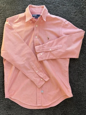 Polo Ralph Lauren Oxfordskjorta Rosa - En riktigt stilig och tidlös rosa oxfordskjorta från Polo Ralph Lauren i fantastisk premiumkvalitet. Den perfekta skjortan för både jobb, fest & vardag! Modellen är 189 cm, men inte så bredaxlad som man kan se. 👕 Märke & Modell: Polo Ralph Lauren - Oxfordskjorta 📏 Storlek & Passform: 15 ½ (L) - Regular fit/ Custom fit 🧵 Material: 100% högkvalitativ bomull 📐 Mått: Bröst (armhåla till armhåla): 60 cm Längd: 80 cm Ärmlängd: 51 cm ✅ Skick: 8,5/10 - Grymt Skick! 💰 Nypris: Ca 1500 kr Skickas inom 24h