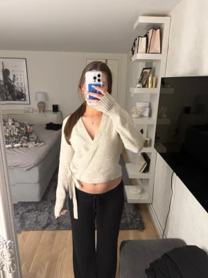 Beige omlottstickad tröja - Mysig beige stickad omlott-tröja med knytband i midjan och långa ärmar. Tröjan har en djup v-ringning och ribbstickade muddar. 