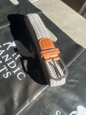 Grått flätat bälte med skinn-detaljer - Snyggt grått flätat bälte med bruna skinn-detaljer och silverfärgat spänne. Perfekt accessoar för att lyfta din outfit med en stilren touch. Bältet har en flexibel design som gör det enkelt att justera. 105cm