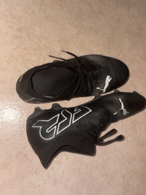 Svarta Puma fotbollsskor med dobbar - Säljer ett par svarta fotbollsskor från Puma med vita detaljer och logga. Skorna har snörning, högre strumpkrage och slitstarka dobbar för bra grepp på planen. Tillverkade i syntetmaterial för lätt känsla och bra bollkontroll.