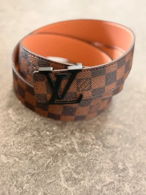 Brunt rutigt LV-bälte i skinn - Snyggt brunt bälte från Louis Vuitton med klassiskt rutigt mönster och stor LV-logga i svart metall som spänne. Bältet är gjort i skinn och har en exklusiv känsla med sina mörka och ljusbruna nyanser. Perfekt accessoar för att lyfta din outfit.