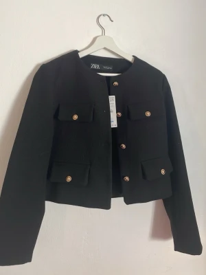 Svart kavaj från ZARA med guldknappar - Stilren svart kavaj från ZARA med rund halsringning och fyra fickor framtill. Kavajen har snygga guldknappar och en rak, kortare passform. Perfekt för en trendig och chic look.