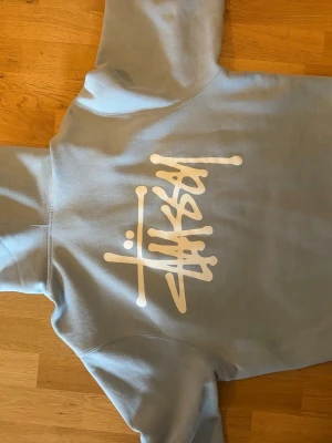 Ljusblå hoodie från Stüssy - Säljer en ljusgrå hoodie från Stüssy med stor vit logga på ryggen och mindre logga på bröstet. Storlek M men Stussy har ganska stor passform så funkar även till L