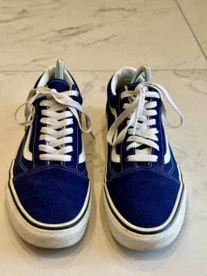 Blå Vans Old Skool sneakers - Klassiska Vans Old Skool sneakers i blått med vit sula och vit sidostripes. Ovandelen är i canvas, med vita skosnören och den ikoniska gummisulan med våffelmönster. Perfekta för en streetwear-look.