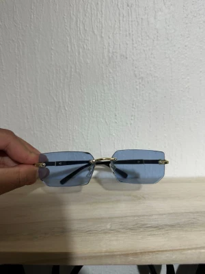 Cart1er glasögon  - Snygga solglasögon med rektangulära, blåtonade glas och guldfärgade detaljer. Glasögonen har minimalistisk design utan synlig båge runt glasen och svarta skalmar. Perfekta för dig som vill ha en modern och stilren look.