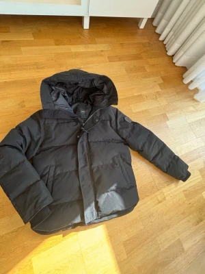 Svart jacka från Canada Goose - Säljer en svart dunjacka från Canada Goose med stor huva och dragkedja framtill. Jackan har två stora fickor och diskret logga på ärmen. Perfekt för kalla vinterdagar tack vare det tjocka dunfodret och den klassiska designen. Storlek M men passar även dig som har storlek S