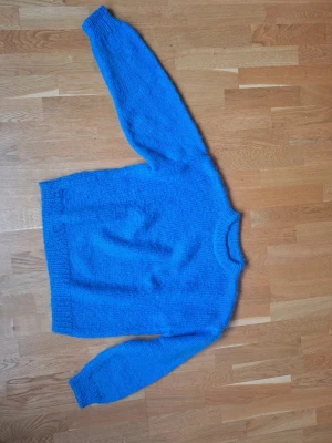 Blå hemstickad tröja i lurvigt garn - En klarblå hemstickad tröja med rund hals och långa ärmar. Tröjan har ribbstickade muddar vid ärmslut och nederkant. Stickat i gosigt sysntetfarn som går bra att tvätta i maskin och ger en mysig känsla.