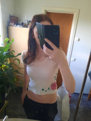 Vit croppad topp med blommönster - Säljer en vit croppad topp med korta ärmar och v-ringning. Toppen har ett fint blommönster i rosa och grönt nere vid sidan, vilket ger en söt och trendig look. Ritat med textilpennor. Perfekt till jeans eller kjol för en fräsch sommarstil.