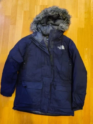Jacka från The North Face  - Jacka från The North Face med huva och fuskpäls. Jackan har lång ärm och klassisk parkasdesign, perfekt för kalla dagar. Logga på ryggen och insidan. Herrjacka. Inga byten.
