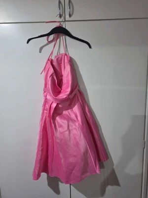 Rosa halterneck klänning med strass - Snygg rosa kortklänning med halterneck och draperad överdel. Klänningen har ett markerat midjeband med strassdetaljer och ett lätt glansigt tyg som ger en festlig känsla. Perfekt för dig som vill sticka ut med en feminin look.