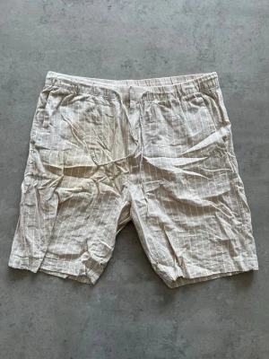 Beige randiga shorts med snörning - L - Säljer ett par beige shorts med tunna vita ränder och snörning i midjan. De har en avslappnad passform och är tillverkade i ett lätt linneliknande material, perfekt för varma dagar. Shortsens design är enkel och stilren med elastisk midja och sidofickor.
