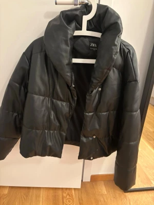 Svart pufferjacka från Zara - Snygg svart pufferjacka från Zara med hög krage och quiltad design. Jackan har en glansig yta som ger en modern look och stängs med tryckknappar framtill. Perfekt för kalla vinterdagar och enkel att matcha till olika outfits.