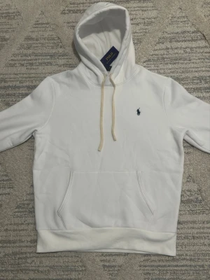 Vit hoodie från Polo Ralph Lauren - Snygg vit hoodie från Polo Ralph Lauren med klassisk blå logga broderad på bröstet. Hoodien har huva med beige dragsko och en stor känguruficka framtill. Tillverkad i mjukt bomullsmaterial och har ribbade muddar vid ärmslut och nederkant.