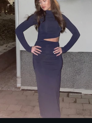 Marinblå långklänning med slits - Snygg marinblå långklänning med lång ärm och hög hals. Klänningen har en trendig cut-out vid midjan och en hög slits på sidan som ger en cool look. Figurnära passform som framhäver siluetten. Perfekt för dig som vill sticka ut.