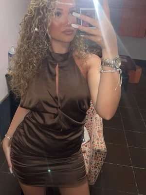 Brun satin kortklänning - Snygg brun kort klänning i satin med hög hals och en liten öppning framtill. Klänningen har en tight passform och är ärmlös, perfekt för dig som vill sticka ut. Materialet ger en lyxig känsla och skimrar snyggt i ljuset.