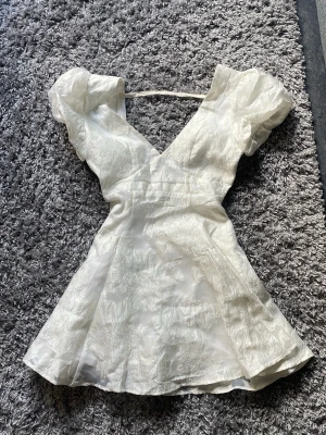 Vit kort klänning med puffärm - Superfin vit kort klänning med v-ringning och puffärmar. Klänningen har ett subtilt blommigt mönster i tyget och är storlek 38. Den passar perfekt för sommaren eller studenten! 