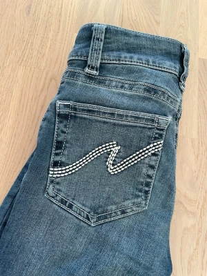 Nelly blå bootcut jeans  - Lite slitet längst ner på jeansen men inget mycket alls! Syns knappt. Snygga blå bootcut jeans med en unik detalj på bakfickan. Jeansen har klassisk femficksdesign, normal midja och utsvängda ben. Perfekta för dig som vill ha en cool och trendig look med lite extra bling.