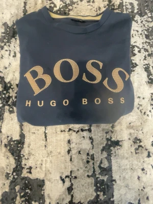 Svart Hugo Boss t-shirt med logga - Snygg svart t-shirt från Hugo Boss med stor beige logga på bröstet. Klassisk rund halsringning och rak passform. Perfekt för dig som gillar stilrena och trendiga plagg med tydlig branding.