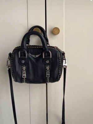 Svart handväska Zadig & Voltaire - Cool svart handväska från Zadig & Voltaire i läder med dubbla handtag och avtagbar axelrem. Väskan har silverfärgade detaljer, två dragkedjor framtill och den ikoniska Zadig & Voltaire-loggan i metall. Perfekt storlek för det viktigaste och ger en edgy vibe.