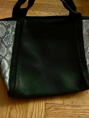 Svart handväska med ormmönster - Snygg svart handväska i konstläder med coola ormmönstrade detaljer på sidorna. Väskan har två rejäla handtag och en praktisk ytterficka med dragkedja. Perfekt för dig som gillar en edgy look och vill ha plats för allt du behöver.