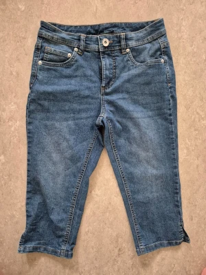 Jeansshorts - Säljer ett par klassiska blå jeansshorts i mjuk denim med fem fickor och knappgylf. Modellen är knälång och har bälteshällor samt snygga söm-detaljer. Perfekta för en avslappnad stil under varmare dagar.