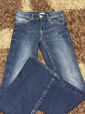 Blå bootcut jeans med broderade fickor - Snygga blå bootcut jeans med slitningar och broderade bakfickor med knappdetaljer. Jeansen har klassisk femficksdesign och kontrastsömmar. Perfekta för dig som gillar en avslappnad men trendig stil.