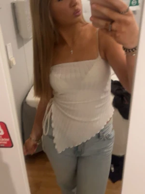 Vit ribbad linnetopp med snörning - Trendig vit linnetopp med smala axelband och snörning i sidan. Toppen är ribbad och har en asymmetrisk nederkant som ger en cool look. Perfekt för varma dagar och enkel att styla med jeans eller kjol.