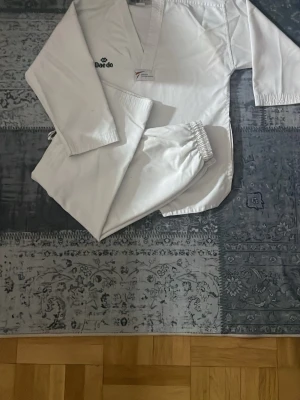 Vit kampsportsdräkt Daedo - Vit kampsportsdräkt från Daedo, perfekt för maskerad eller cosplay. Setet består av en långärmad överdel med V-ringning och byxor med resår i midjan. Tillverkad i slitstarkt bomullstyg och har Daedo-logga på bröstet. Enkel och autentisk design för kampsportskaraktärer.