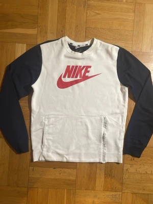 Nike sweatshirt med svarta ärmar - Säljer en snygg vit Nike sweatshirt med svarta ärmar och stor röd Nike-logga på bröstet. Tröjan har en stor magficka med dragkedja på ena sidan där det står Nike på bandet. Tillverkad i mjukt bomullsmaterial, perfekt för chill dagar.