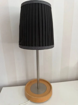 IKEA Bäve Bordslampa med svart skärm - Snygg bordslampa med cylinderformad svart textilskärm med veckad design och grå kant. Lampfoten är i ljust trä och metall, vilket ger en modern och stilren känsla. Perfekt som belysning på skrivbord eller nattduksbord.