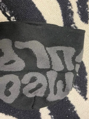 Svart och grå mössa med text - Säljer en svart mössa med grå text i stor, grafisk stil runt om. Mössan är stickad i mjukt material och har en avslappnad passform. Perfekt för dig som gillar streetwear och vill ha en enkel men snygg accessoar till hösten och vintern.