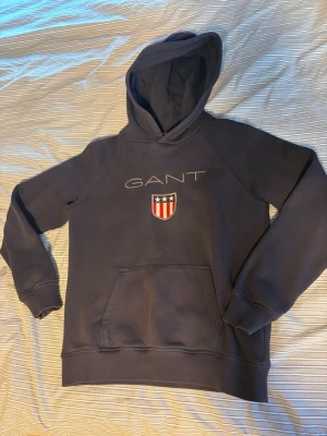 Mörkblå hoodie från GANT - Snygg mörkblå hoodie från GANT med broderad logga och ett sköldmärke på bröstet. Klassisk känguruficka framtill och stor huva. Tillverkad i mjukt bomullsmaterial som känns skönt mot huden. Perfekt för en avslappnad och stilren look.