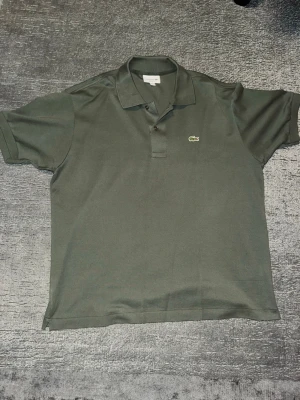 Mörkgrön pikétröja från Lacoste - Klassisk mörkgrön pikétröja från Lacoste med den ikoniska broderade krokodilloggan på bröstet. Tröjan har korta ärmar, krage och knappar framtill. Perfekt för en clean och sportig look. Äkta!! Qr kod skannar
